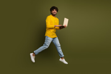 Young man in casual attire enthusiastically using a laptop while leaping against a moss green...