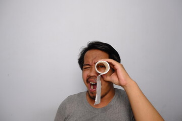 Amused man in a gray shirt using a paper roll as a pretend telescope while making a funny facial...