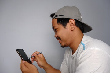 Smiling Asian man in a white t-shirt and gray cap using a stylus on a digital tablet, captured in a...