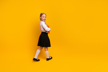 Charming schoolgirl in uniform confidently walking with book against a vibrant yellow background,...