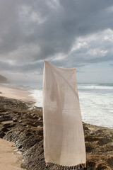 A white textile towel stands on a rocky beach, catching wind under overcast skies. Natural textures...