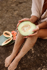 A woman holds a halved melon while seated on the rocks, surrounded by tropical fruits. Slow island life.