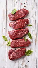 Fresh raw beef steaks with marbling arranged artfully on rustic white wooden surface, garnished with green basil leaves and scattered peppercorns