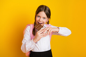 Smiling schoolgirl holding chocolate bar in hands standing against vibrant yellow background wearing uniform with braids