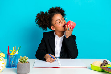 Young schoolgirl in formal attire sitting at a desk with supplies eating a red apple against...