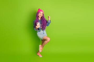 Cheerful young woman with vibrant purple hair using phone and celebrating on a fresh green background, showcasing modern style