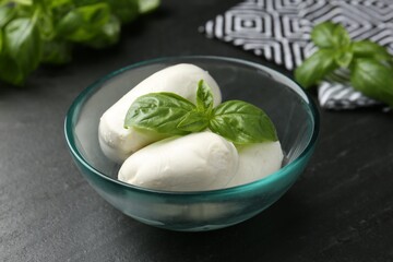 Pieces of delicious mozzarella cheese and basil in bowl on dark textured table, closeup