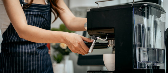 A young Asian female barista in a striped apron carefully pours steamed milk to create intricate...