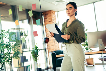 Confident young female professional holding folder, standing in modern office amid colorful notes,...