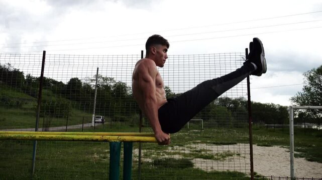 Strong man trains abdominal muscles raising legs on parallel bars at sports ground. Young shirtless sportsman exercising at nature. Sporty athlete working out at nature. Concept of sportive lifestyle