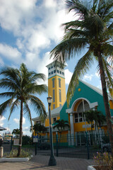 Tall yellow and white clock tower with louvered windows, framed by palm leaves, under a bright blue...