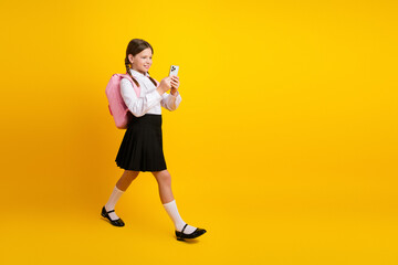 Happy schoolgirl in a uniform with backpack using smartphone on a yellow background