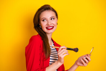 Obraz premium Charming woman holding a brush and mirror smiling with vibrant yellow background, wearing stylish red outfit, concept of beauty