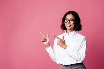 Charming young woman with stylish formalwear pointing sideways on a vibrant pink background,...