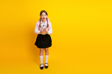 Excited schoolgirl with a smartphone in formal uniform on a yellow background, expressing...