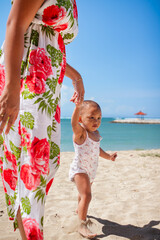 Asian mother in floral dress holding the hand of her toddler girl, helping her walking barefeet on beach sand under harsh sunlight on hot sunny summer day with blue ocean and blue sky on background