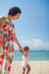 Asian mother in floral dress holding the hand of her toddler girl, helping her walking barefeet on beach sand under harsh sunlight on hot sunny summer day with blue ocean and blue sky on background