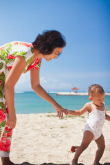 Asian mother in floral dress holding the hand of her toddler girl, helping her walking barefeet on beach sand under harsh sunlight on hot sunny summer day with blue ocean and blue sky on background