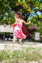 Cheerful Asian little girl in colorful casual tropical dress running and jumping around on the beach with a big smile on her face