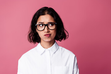 Confident young woman posing against pink backdrop wearing glasses and white formal shirt exuding...