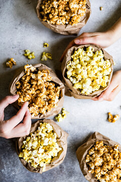 An assortment of popcorn flavors in brown paper bags, with hands reaching for the snacks on a textured surface.