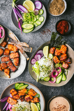 An overhead shot with a custom gray stone textured surface with two beautiful ceramic bowls filled with rice, fresh vegetables, seaweed, and crispy, golden brown pieces of salmon. On the table is a pl