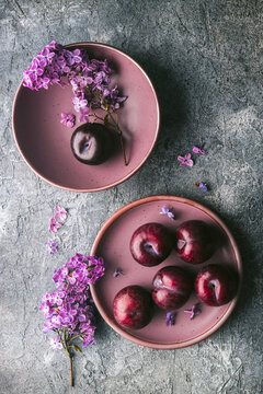 Two mauve-colored bowls on a custom grey, textured surface with an arrangement of several ripe plums. Lilac flower branches are artfully arranged creating a soft, pleasing image.