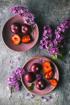 Two mauve-colored bowls on a custom grey, textured surface with an arrangement of several ripe plums, one cut in half, one in thirds to reveal the flesh. Lilac flower branches are artfully arranged cr