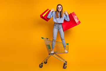Young woman in stylish blue shirt holding shopping bags on a shopping cart with orange background