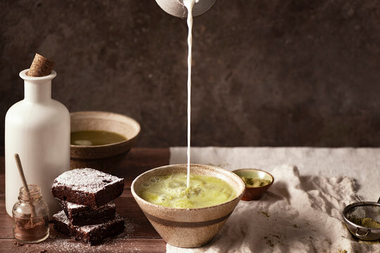 two bowls of matcha tea with brownies with cocoa powder on table, milk being poured in the bowl