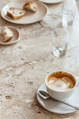 A cappuccino in a white ceramic cup on a marble table, with a spoon on the saucer. Blurred plates with bread and clear water glasses in the background create a calm café scene.