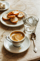 A cup of espresso served on a saucer with a silver spoon, next to a glass of iced water and a plate of sliced baguette. Captured on a marble table in a softly lit café setting.
