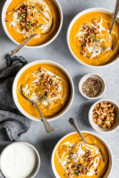 Five bowls of creamy pumpkin soup garnished with nuts and herbs, surrounded by small bowls of spices and nuts on a textured grey surface.