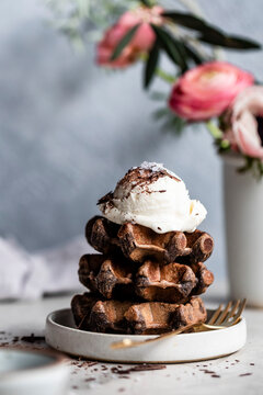A stack of chocolate waffles topped with a scoop of vanilla ice cream and chocolate shavings, placed on a white plate with a fork nearby, with pink flowers blurred in the background.