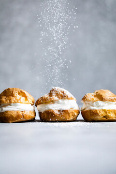Three cream puffs being sprinkled with powdered sugar on a gray background.