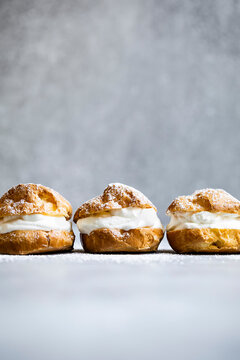 Three cream puffs filled with whipped cream are arranged in a row on a light background, dusted with powdered sugar.
