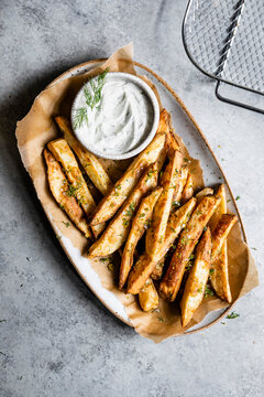 A plate of crispy potato wedges garnished with herbs, served with a bowl of creamy dipping sauce.