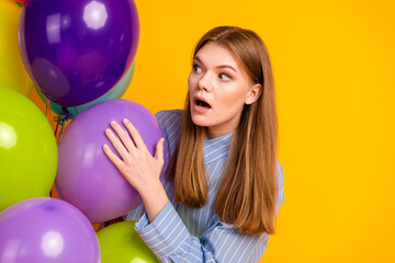 Surprised young woman holding colorful balloons against a vibrant yellow background, dressed in a blue striped shirt