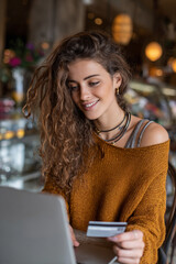 Woman with curly hair holding a credit card while looking at a laptop in a cafe setting indoors