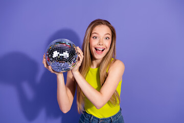 Excited young woman holding a sparkling disco ball against a vibrant purple background exuding...