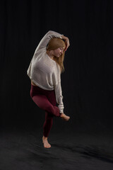 a person performing a yoga pose, balancing on one foot while holding the other with an opposite hand. They are dressed in a light-colored sweater and maroon leggings