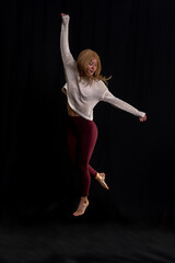 a person performing a yoga pose, balancing on one foot while holding the other with an opposite hand. They are dressed in a light-colored sweater and maroon leggings, standing against a dark backgroun