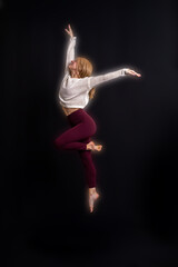 captures a dancer in a graceful pose on a black box against a dark backdrop. Dressed in a light gray sweater and maroon leggings, the dancer extends their arms upward while maintaining balance with on