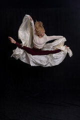 captures a dancer in a graceful pose on a black box against a dark backdrop. Dressed in a light gray sweater and maroon leggings, the dancer extends their arms upward while maintaining balance with on