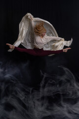 captures a dancer in a graceful pose on a black box against a dark backdrop. Dressed in a light gray sweater and maroon leggings, the dancer extends their arms upward while maintaining balance with on