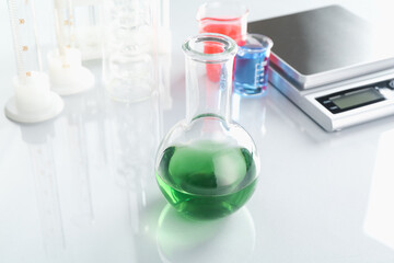 Flask with green liquid, other laboratory glassware and scale on white mirror table, closeup