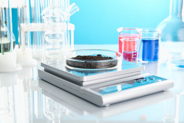 Scale with Petri dish of sample and other laboratory glassware on white mirror table against light blue background, closeup