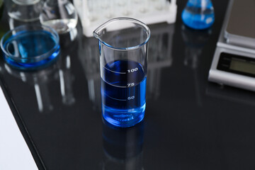 Beaker with blue liquid, other laboratory glassware and scale on black mirror table, closeup
