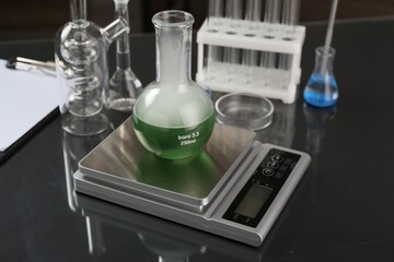 Scale with flask of sample and other laboratory glassware on black mirror table, closeup