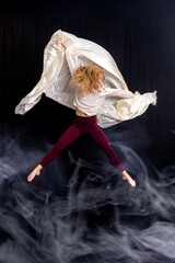 captures a dancer in a graceful pose on a black box against a dark backdrop. Dressed in a light gray sweater and maroon leggings, the dancer extends their arms upward while maintaining balance with on